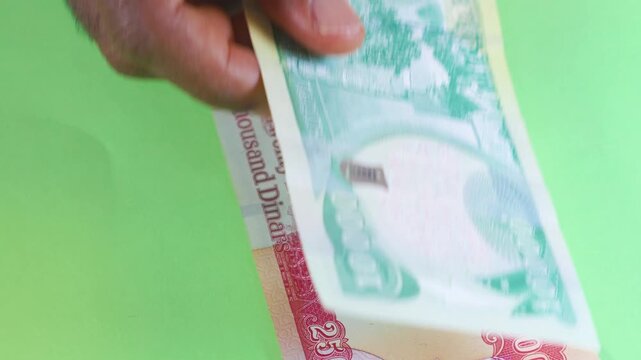 A hand places several Iraqi Dinar banknotes on a green background. The video displays various denominations, highlighting the colorful artwork and official markings of Iraq's national currency.