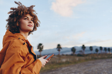 Young woman with curly hair wearing orange jacket smiles holding smartphone outdoors, lifestyle...