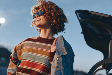 Smiling woman with curly hair wearing a rainbow sweater and sunglasses holding a denim jacket near...