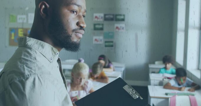 Male teacher reading clipboard, turning forward and monitoring students writing on paper in class