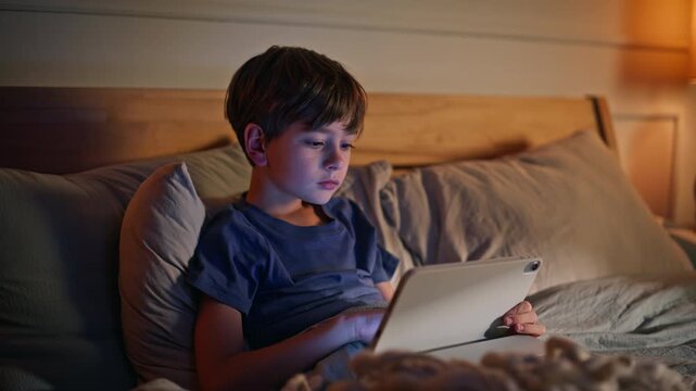 Boy browsing tablet night bedroom closeup. Little kid focused computer lying bed