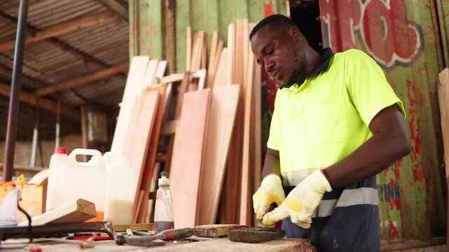 African carpenter, wearing safety gloves, sharpens a blade with a traditional sharpening stone on a rustic wooden workbench in his workshop, closeup