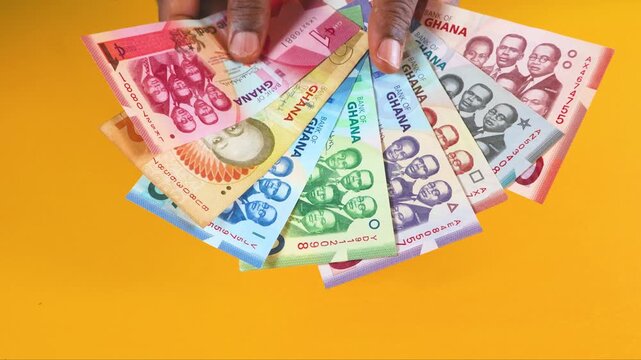 A person fans out a colorful collection of Ghana Cedi banknotes against a bright yellow backdrop. The video highlights various denominations, showing the detailed portraits and vibrant national art.