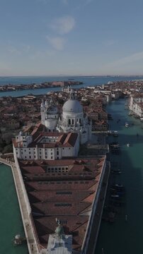 Vertical drone shot ascending from Punta della Dogana in Venice showing Basilica Santa Maria della Salute, canal, boats, and cityscape in the background.