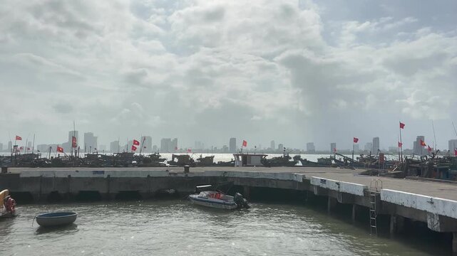 Fishing boats gathered along concrete pier at Son Tra Marina in Da Nang with red flags raised above decks while small motorboat moves across harbor water beneath cloudy daylight sky