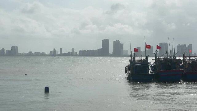 Fishing boats anchored near Son Tra Marina in Da Nang with red flags raised above decks while calm harbor water reflects light and distant city skyline appears beneath cloudy sky
