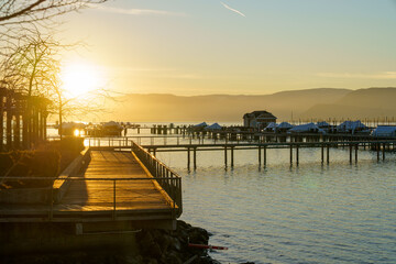 Fototapeta premium A warm golden sunset casts light across a calm lakeside marina, illuminating wooden docks and boat slips. The sun reflects off the water while distant mountains form soft silhouettes along the horizon