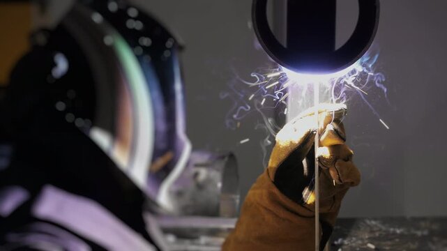Industrial worker performing manual arc welding with electrode and bright sparks during steel fabrication process in heavy manufacturing factory.