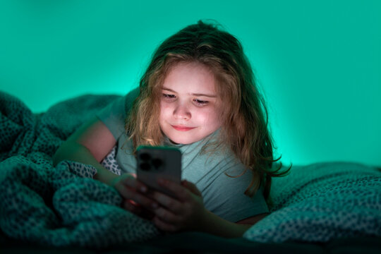 Teenager chatting on phone in bed at night. Child texting on phone in bed at night. Kid scrolling phone under blanket at night. Teenager browsing internet on phone in bed at night.