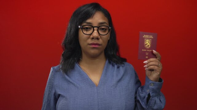 African american woman wearing glasses holding finland passport in studio against red wall; confidence identity travel.