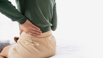 Woman holding her lower back in pain while sitting on bed. Representing back pain, muscle strain, spine problem and healthcare or body pain concept.