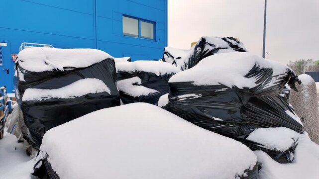Cargo in black foil stored in front of blue warehouse under thick layer of snow, winter logistics and outdoor storage, industrial goods protected against frost and precipitation.