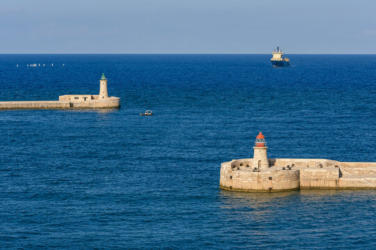 Faros gemelos se&ntilde;alando la entrada al puerto de La Valeta, Malta, con un buque de carga navegando en el horizonte. Seguridad mar&iacute;tima.