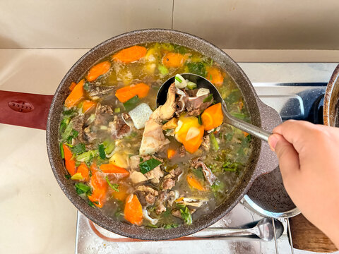 Indonesian beef cartilage soup or sup daging tulang rawan with carrots and potatoes simmering in rich broth. Woman hand stirring homemade traditional soup in cooking pot on kitchen stove.