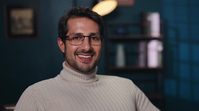 Portrait of man in glasses smiles to camera in living room interior. Bearded guy in turtle neck sweater expresses positive mood in domestic environment