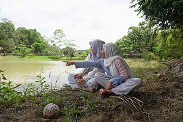 Fototapeta premium Two happy young Asian Muslim sisters sitting under a tree by the lake at sunset, chatting and laughing together