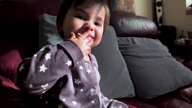 Childhood nutrition and self-feeding a cute South Asian baby girl is eating food with her hands on a sofa at home exploring textures and tastes during early development and independence