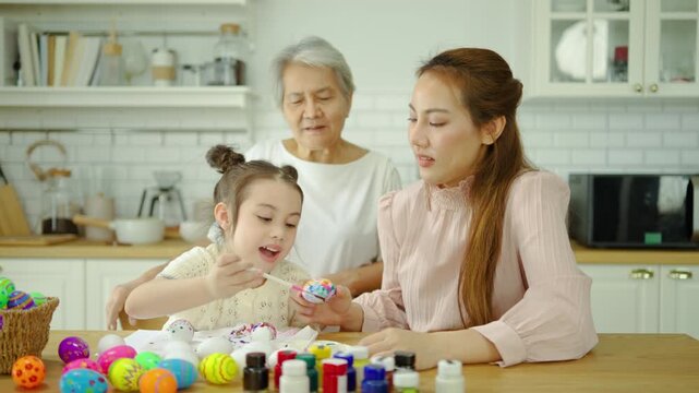 Happy three-generation Asian family, grandmother, mother, and daughter, painting colorful Easter eggs together in a bright kitchen. A joyful holiday tradition and family bonding activity at home.