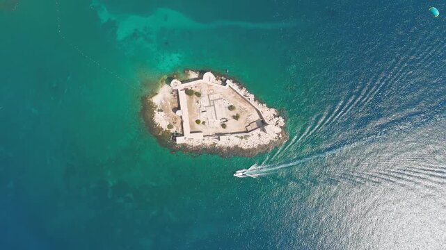 Kizkalesi, Mersin, Turkey. Dynamic parasail flight over Maiden Castle island fortress, surrounded by waves and rocks, towed by speedboat.. Aerial View, HEAD OVER SHOT with rotation