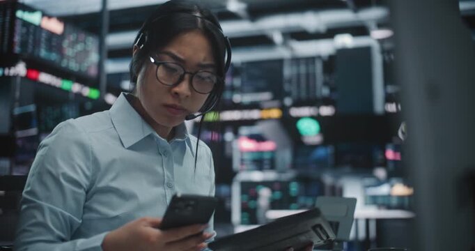 Asian Woman Frantically Cross References Real Time Financial Data on Multi Monitor Workstation and Tablet. Female Reports Critical Market Information Contact on Smartphone. Urgent Communication.