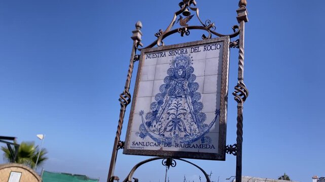 Ornate wrought iron sign displaying a blue and white azulejo tile depicting Nuestra Senora del Rocio against a clear sky