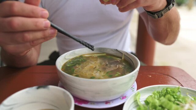 Caucasian Traveler Squeezing Lime Over Vietnamese Pho Bo Bowl, Closeup Hands Adding Citrus To Broth, Visible Beef Slices And Noodles, Side Plate Of Greens, Tangy Seasoning Moment, Outdoor