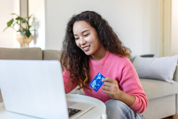Fototapeta premium Happy Asian woman using credit card for online shopping on laptop at home. Female buyer making digital payment or banking. E-commerce, finance and consumerism concept in modern bright apartment.
