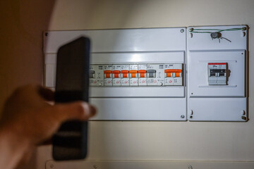 Person's hand holding a smartphone, using its flashlight to illuminate an electric fuse box with multiple circuit breakers, solving an electrical problem during a power cut