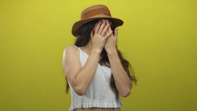Young hispanic woman wearing straw hat pinches nose with both hands in studio against yellow backdrop; disgust.