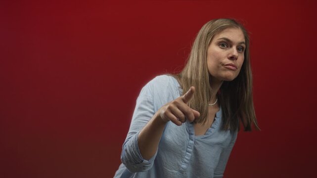 Young blonde woman points finger at camera in studio with red wall, mouth open and direct gaze; surprise accusation.