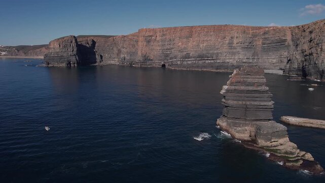 Stratified sea stack rising from Atlantic Ocean below towering fal&eacute;sias of Vila Nova de Mil Fontes Alentejo Portugal showing sedimentary rock layer and marine erosion pattern, drone low pull away