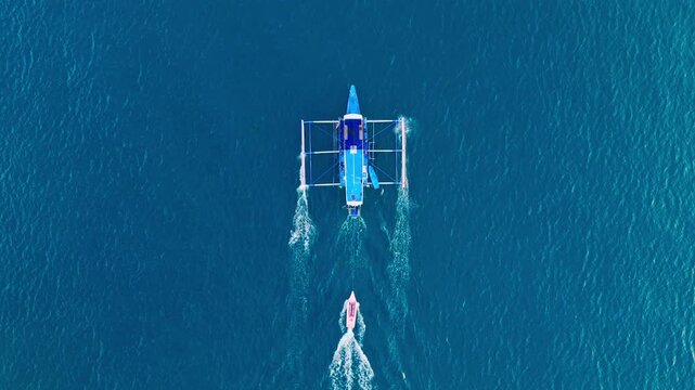 Top down drone view of large outrigger towing smaller pink boat across ocean