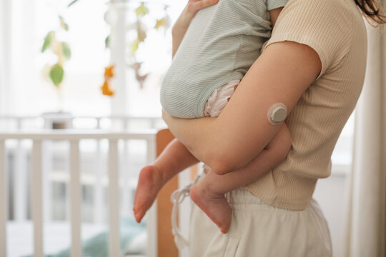 Close-up of a woman in light homewear with a Continuous Glucose Monitoring on her arm holding a baby next to a white crib with a hanging musical mobile toy. 
