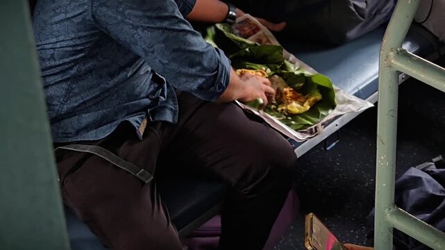 A man in a blue shirt enjoys a traditional meal served on a banana leaf while traveling by train. The authentic experience of Indian railways, featuring blue berths & the bustle of a journey. No face.