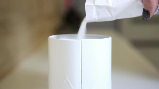 Detail view of white powdery substance gently falling from small paper packet into clean white cylindrical vessel, action suggests meticulous cooking or beverage preparation