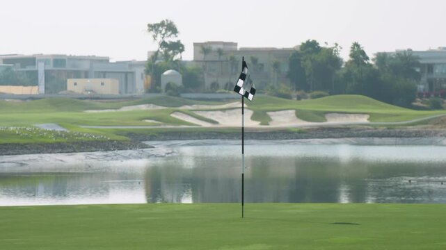 Golf course scene with a checkered flag near a serene lake, showcasing lush green grass and modern buildings in the background