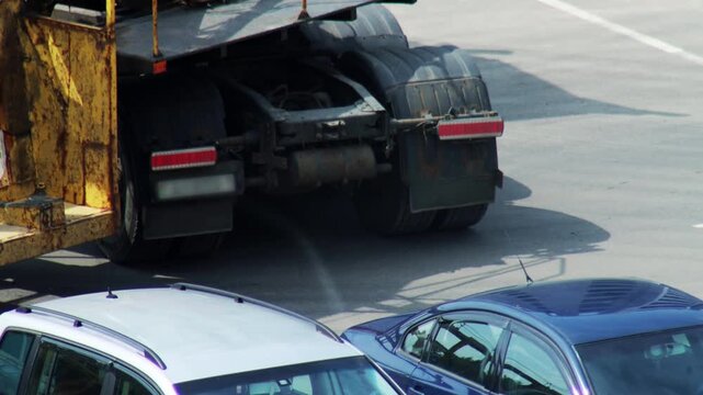 High angle view of rear section of large yellow heavy duty truck on asphalt surface, brake lights of truck illuminate, two passenger vehicles pass on sunny day, logistics and urban mobility
