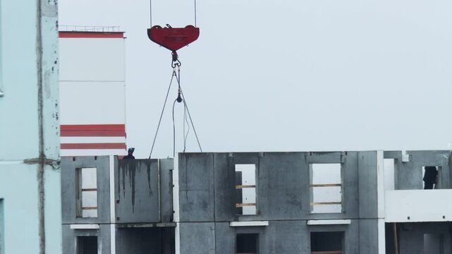 Heavy industrial crane hook with chains suspends precast concrete wall panel for building assembly. Worker on framework guides panel placement. Urban development.