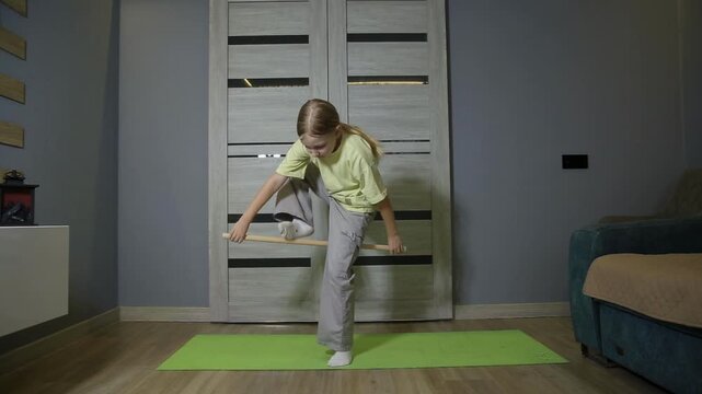 Young girl engaged in active leg workout during home fitness routine. She stands on a green yoga mat, holding a wooden stick horizontally with both hands for balance.