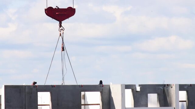 Close up view of industrial red crane hook and robust chain assembly carefully positioning a large grey precast concrete wall panel for new building construction.