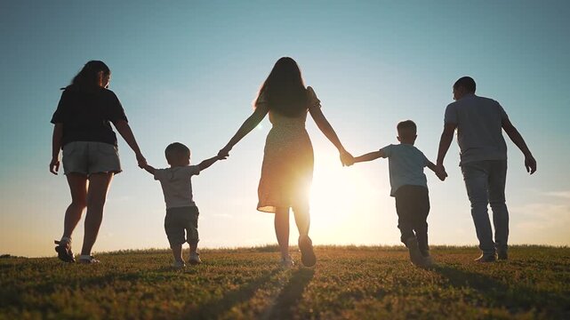 Big happy family is walking in green field at sunset. Parents and children hold hands in a row. Group of people enjoys summer evening in nature. Large family walks away together in nature park happily