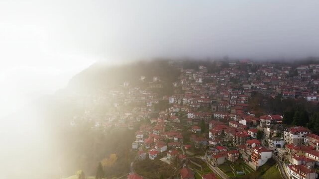 Metsovo hillside village with red roofs and heavy fog in Epirus Greece, sunlight diffuses over edge, aerial