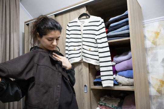 Young woman holding up a brown corduroy shirt to her shoulders while looking at her reflection near an open wardrobe