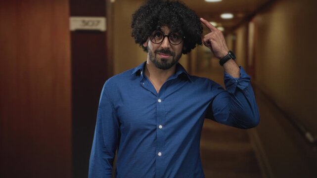 Man wearing glasses and blue shirt points finger to head in hotel corridor near room sign, beard visible; thinking thoughtful curiosity.