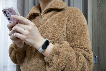 Close-up of a young woman's hands holding a mobile phone, wearing a brown teddy jacket and a fitness tracker © Iuliana