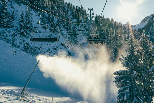 Skiers ride chairlift over snow gun on sunny day at Whistler