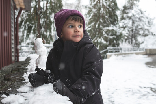 Young boy playing in snow leans up on wall and looks at snowflakes