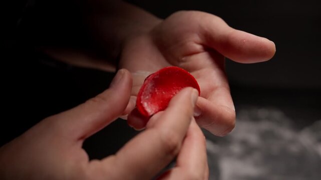Close-up hands carefully shaping delicate red dumpling wrapper with savory filling, showcasing precise folding technique and craftsmanship in handmade cooking process