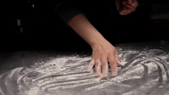 Slow motion hands dusted with flour preparing a dark kitchen countertop for baking, smoothing and clapping the powdery surface in dramatic low key lighting