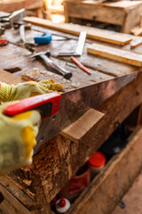 African male carpenter in work gloves saws a wooden plank with a hand saw on a rustic workbench in his workshop